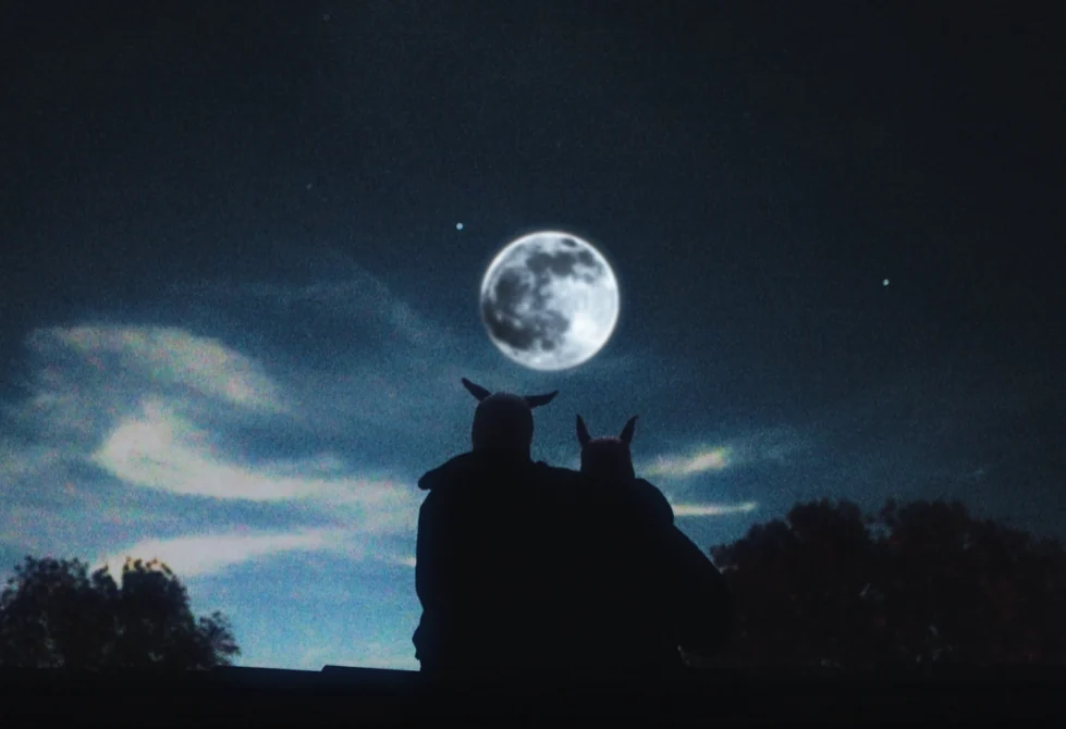 Two silhouetted figures wearing animal ear hoods sit beneath a full moon in a night sky