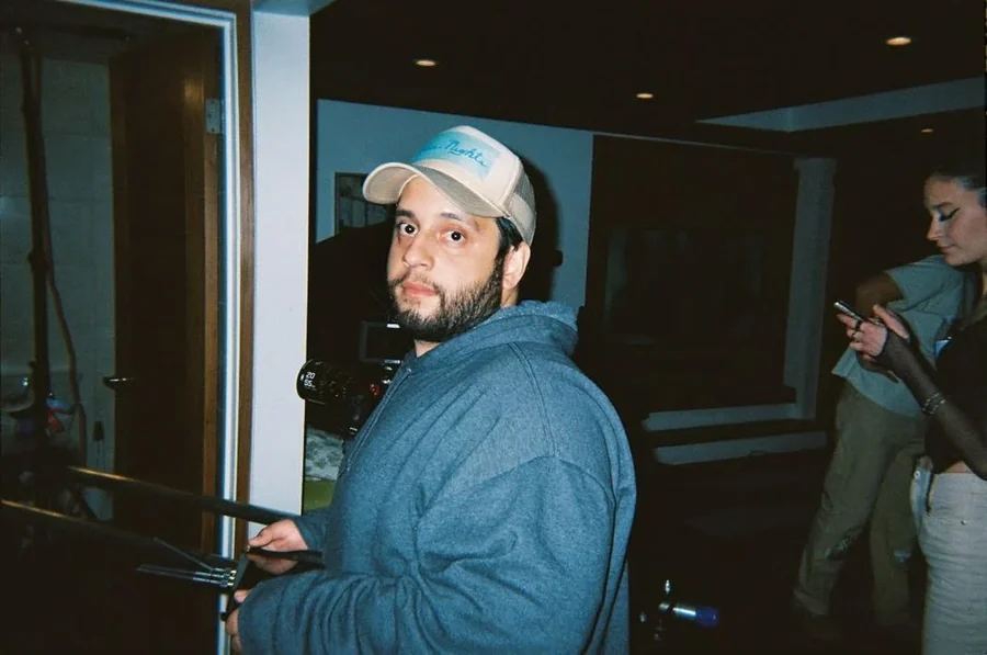 Marcos Segura in a cap and blue hoodie holding a camera rig on set, looking toward the lens